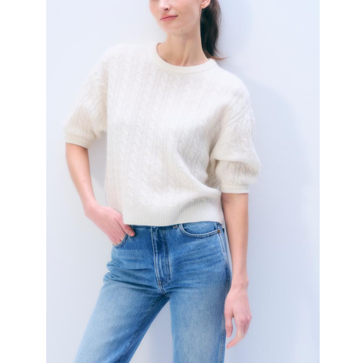 A woman wearing a cream-colored textured short-sleeve knit sweater and blue jeans, standing against a light blue-gray background.