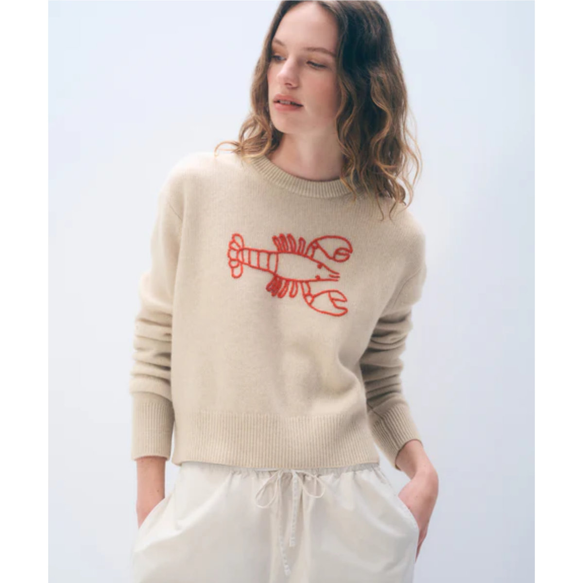 A woman wearing a beige knit sweater with a red lobster graphic on the chest, paired with white drawstring pants, against a light gray studio background.