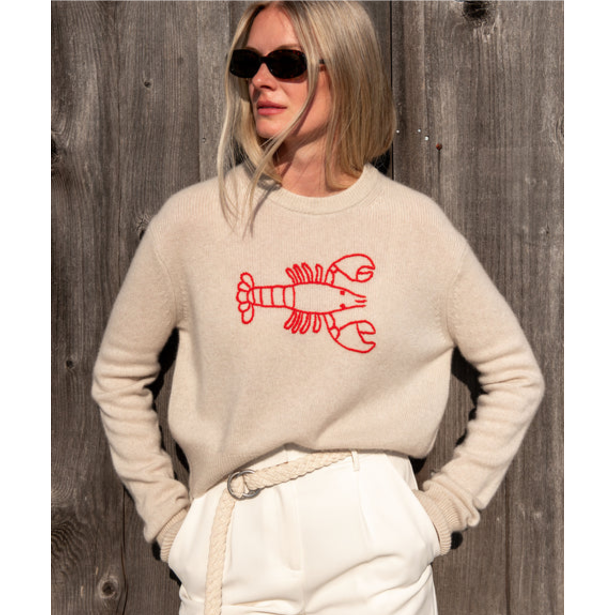 A woman wearing a cream-colored crewneck sweater with a red embroidered lobster graphic on the chest, paired with white trousers and sunglasses, standing against a rustic wooden wall.