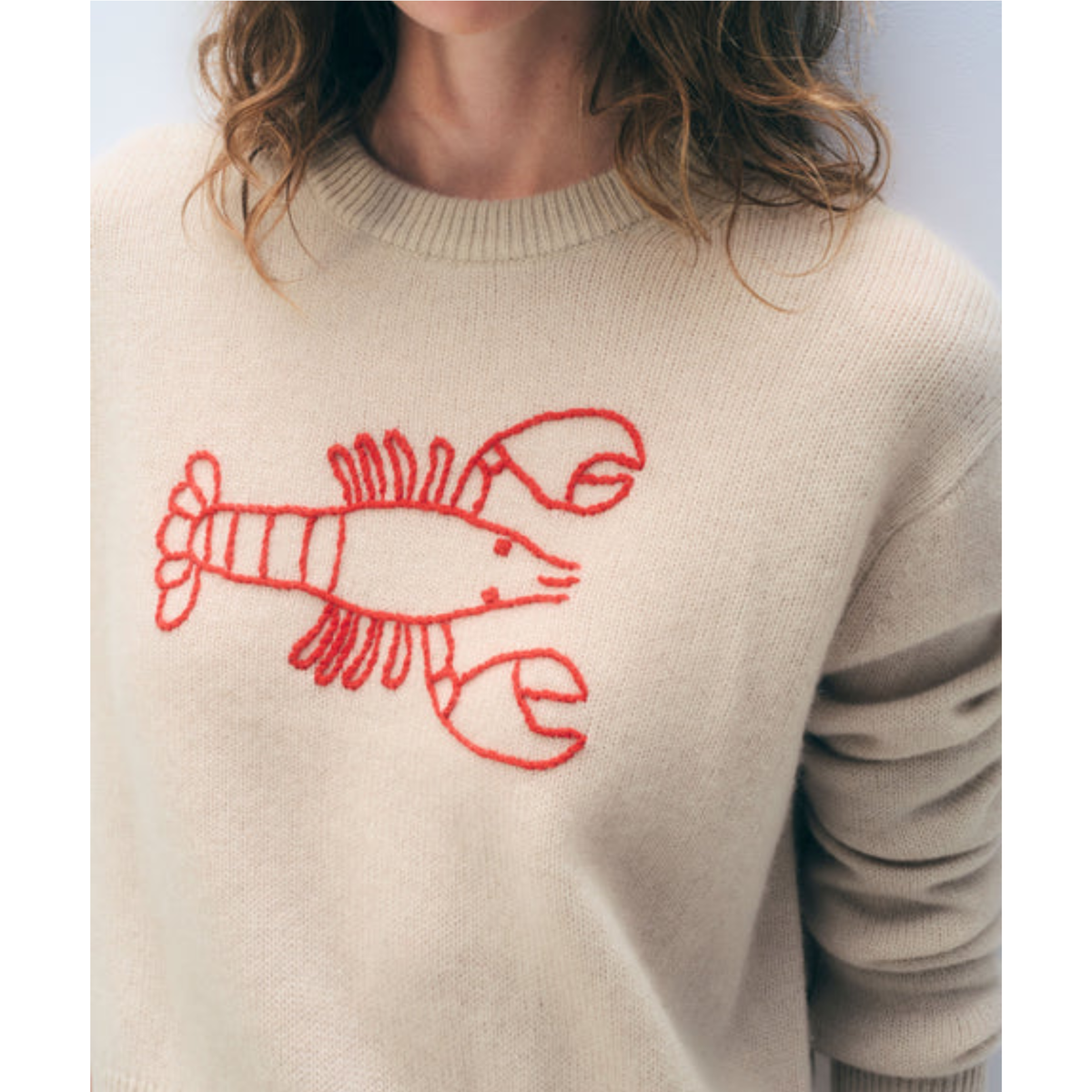 A close-up of a person wearing a cream-colored knit sweater with a red embroidered lobster graphic on the chest.