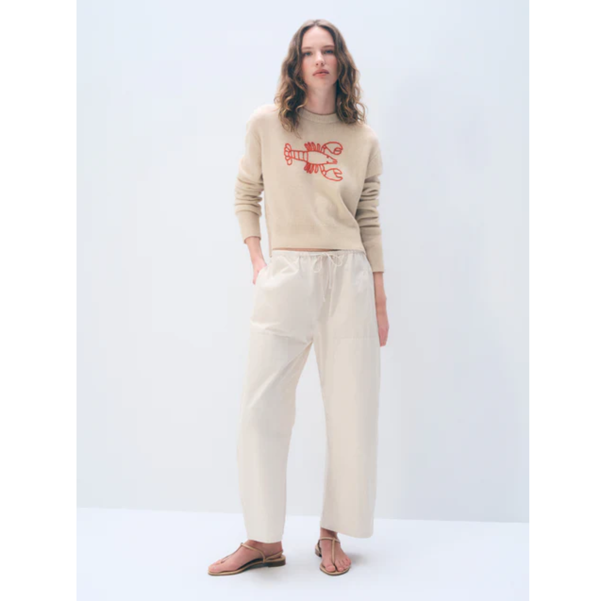 A woman modeling a beige crewneck sweatshirt with a red graphic print and matching beige wide-leg trousers, standing against a light gray studio background.