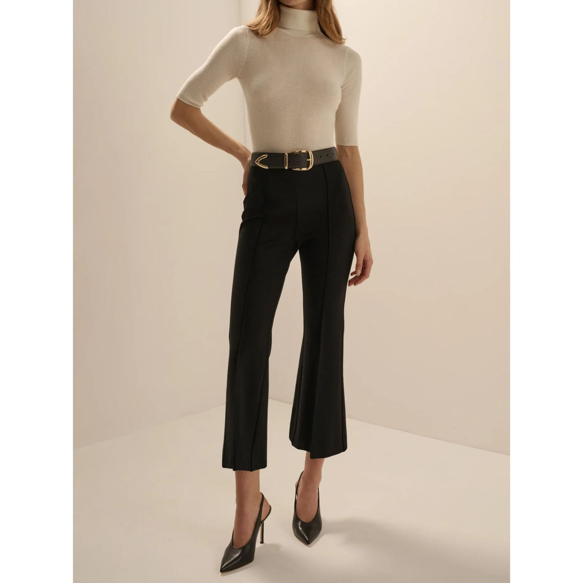 A model wearing black cropped flared trousers with a gold buckle belt, paired with a cream short-sleeve mock-neck top and black pointed-toe heels, set against a warm beige studio background.