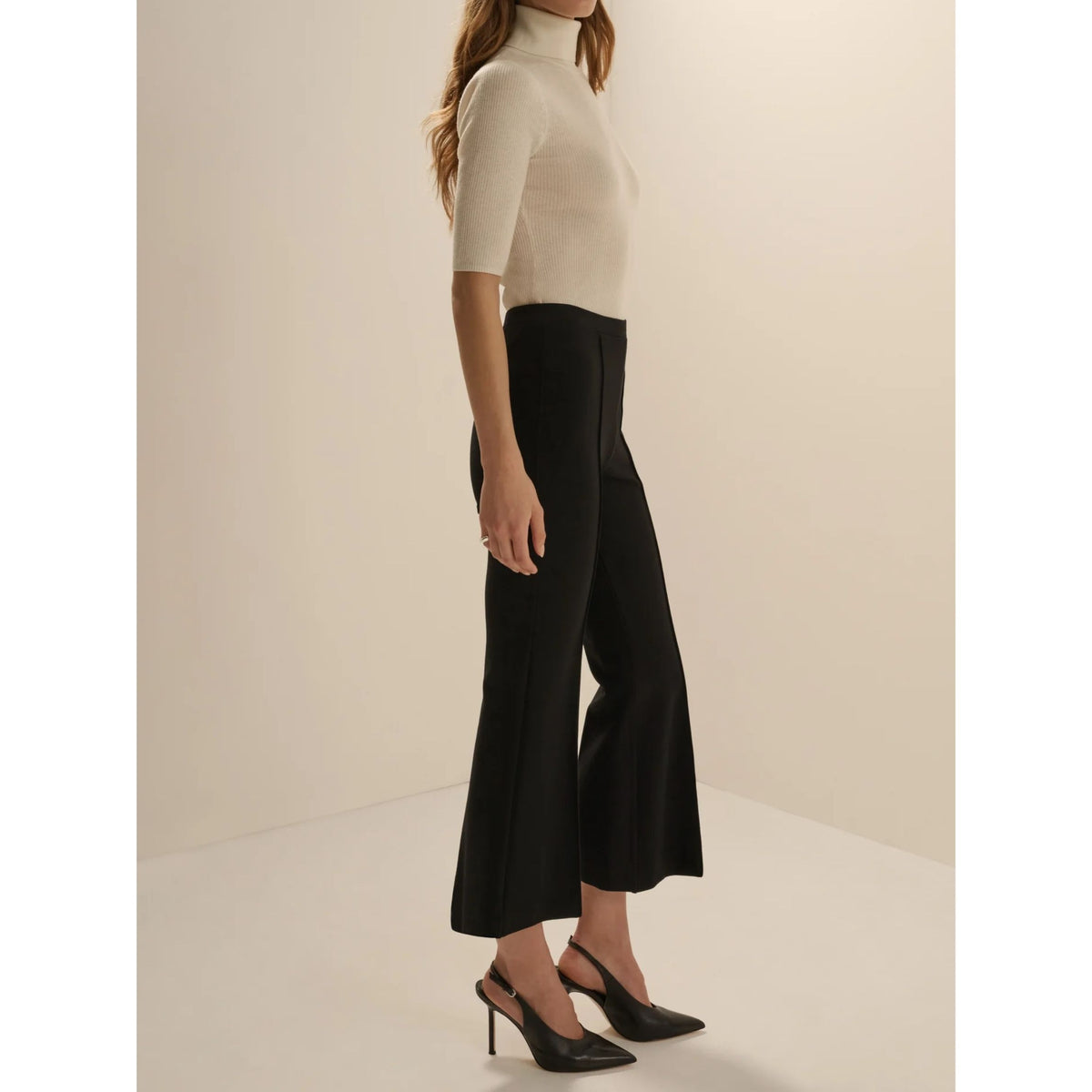 A woman modeling black wide-leg cropped pants with a high waist, paired with a cream short-sleeve mock-neck top and black slingback heels, set against a minimalist beige studio background.