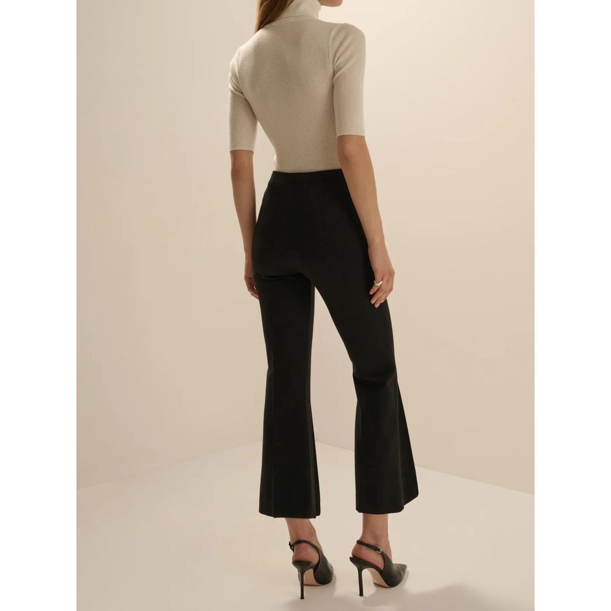 A model shown from the back wearing black flared cropped pants and a beige short-sleeve mock-neck top, paired with black high-heeled sandals, set against a plain beige studio background.