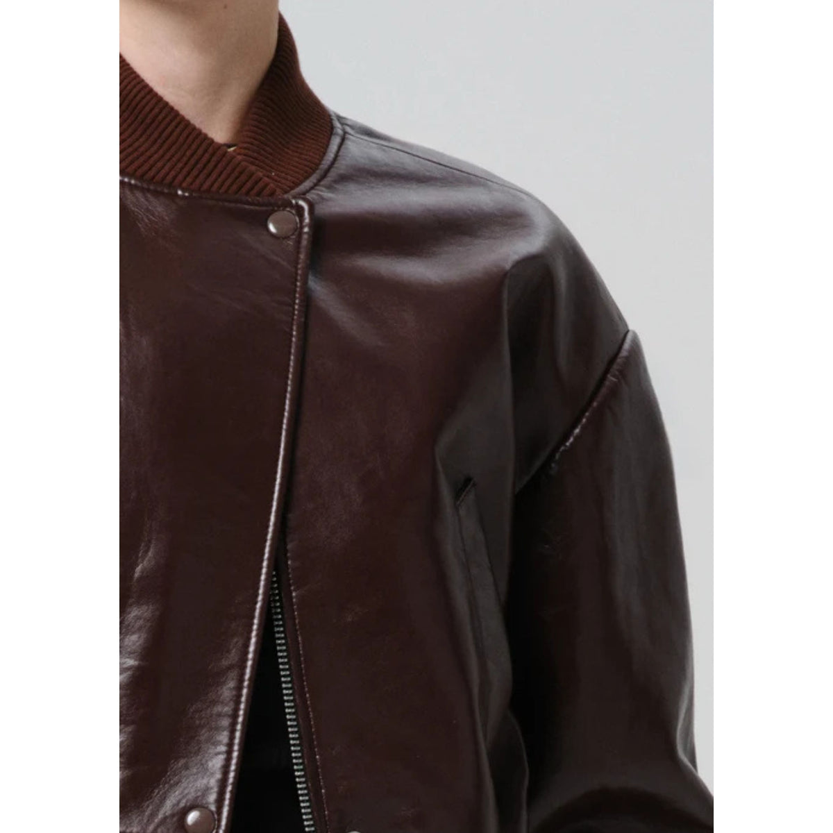 Close-up of a dark brown leather jacket with a ribbed brown knit collar, featuring a front zipper and metal snap button, worn by a model against a light gray background.