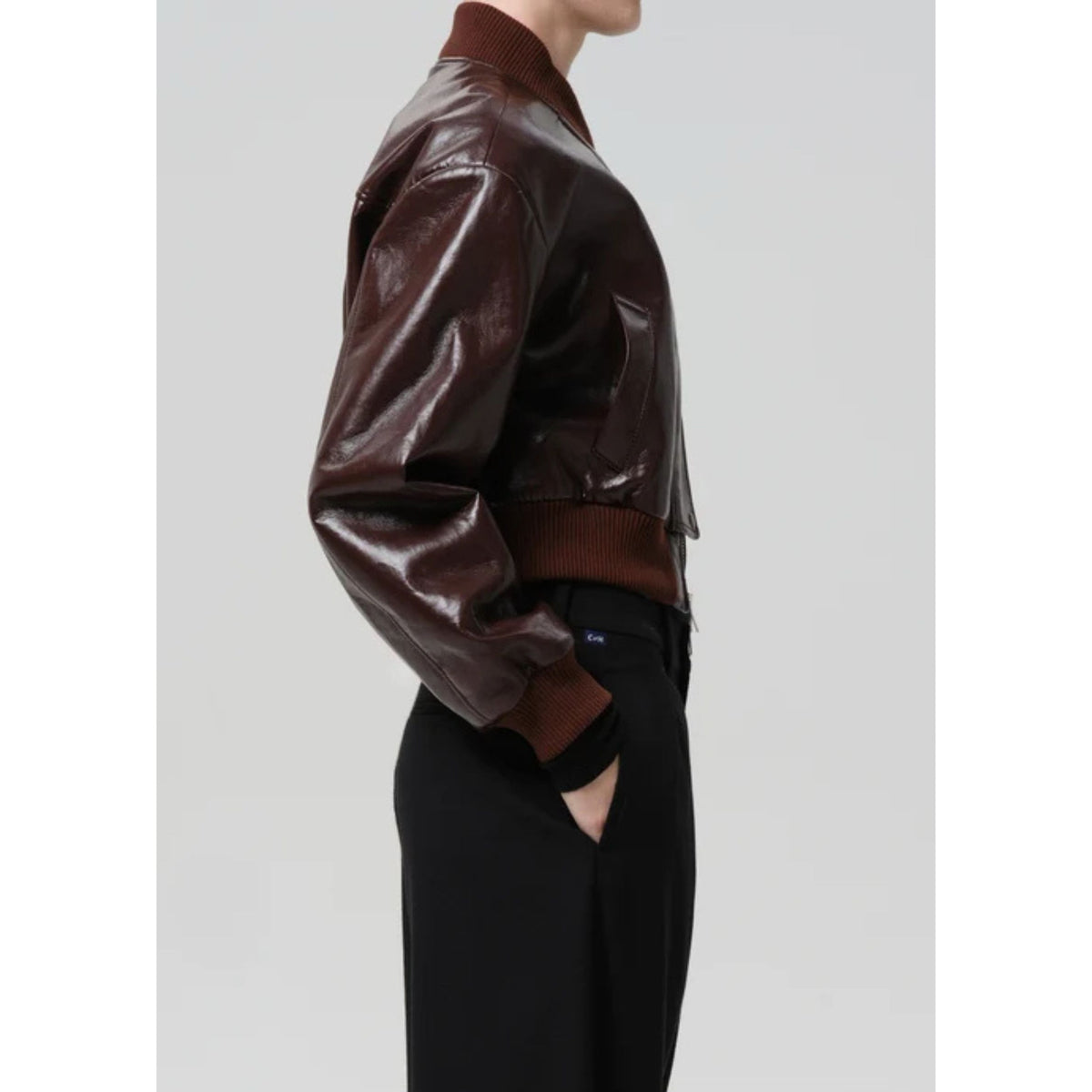 A side-profile view of a person wearing a glossy dark brown faux-leather bomber jacket with ribbed brown knit collar, cuffs, and hem, paired with black pants, against a light gray studio background.