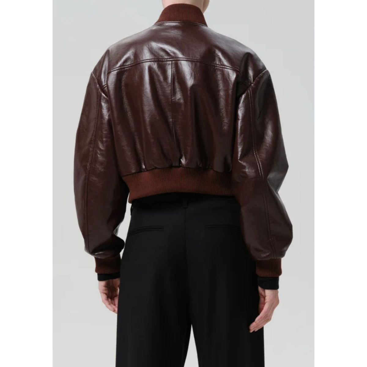 A person shown from the back wearing a cropped dark brown leather bomber jacket with ribbed collar, cuffs, and hem, paired with black pants, against a light gray studio background.