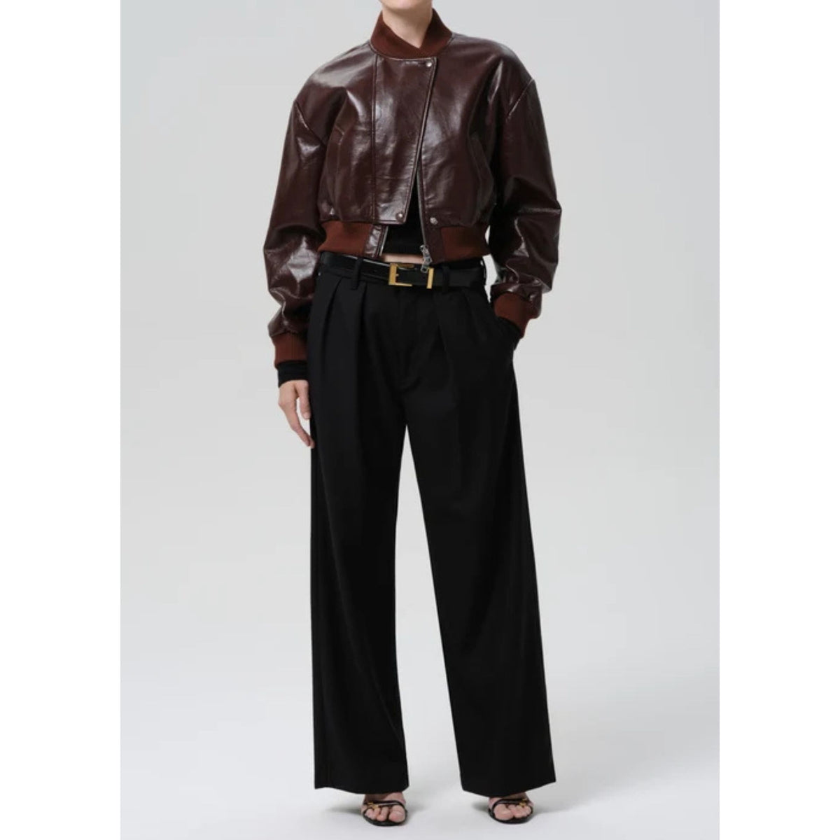 A model wearing a dark brown faux-leather bomber jacket, black wide-leg trousers, and a black belt with a gold buckle, standing against a light gray studio background.