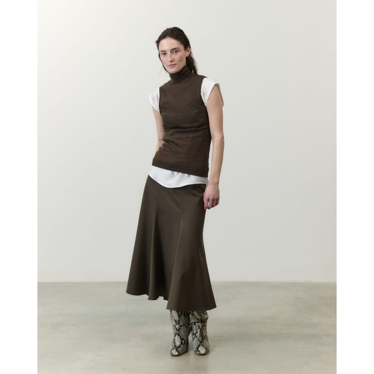 A model stands in a minimalist studio setting wearing a dark brown sleeveless mock-neck top over a white turtleneck, paired with a matching dark brown midi skirt and snakeskin-patterned boots.