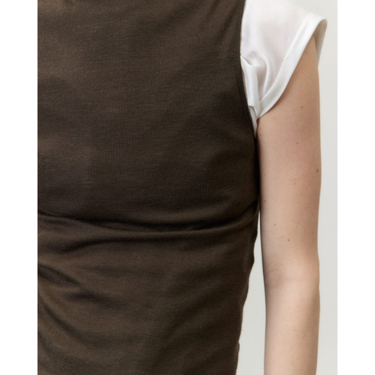 Close-up of a person wearing a sleeveless dark brown knit top or dress layered over a white short-sleeve top, against a plain light background.