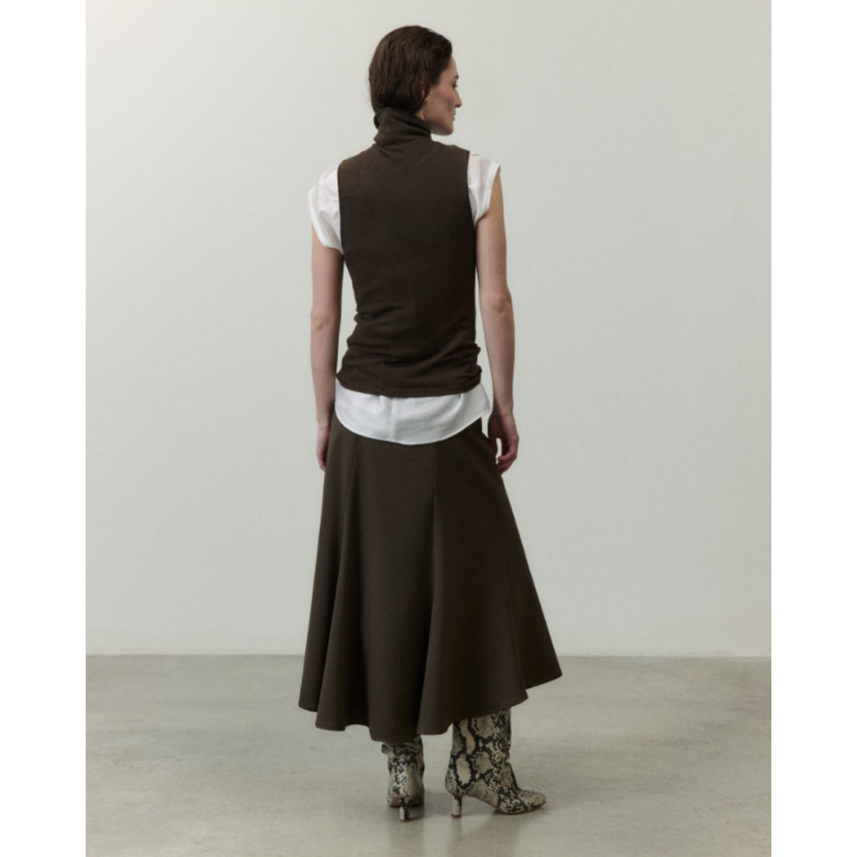 A model shown from the back wearing a dark brown sleeveless turtleneck vest over a white short-sleeve top, a matching dark brown midi skirt, and snakeskin-patterned heeled boots, set against a minimalist studio background.