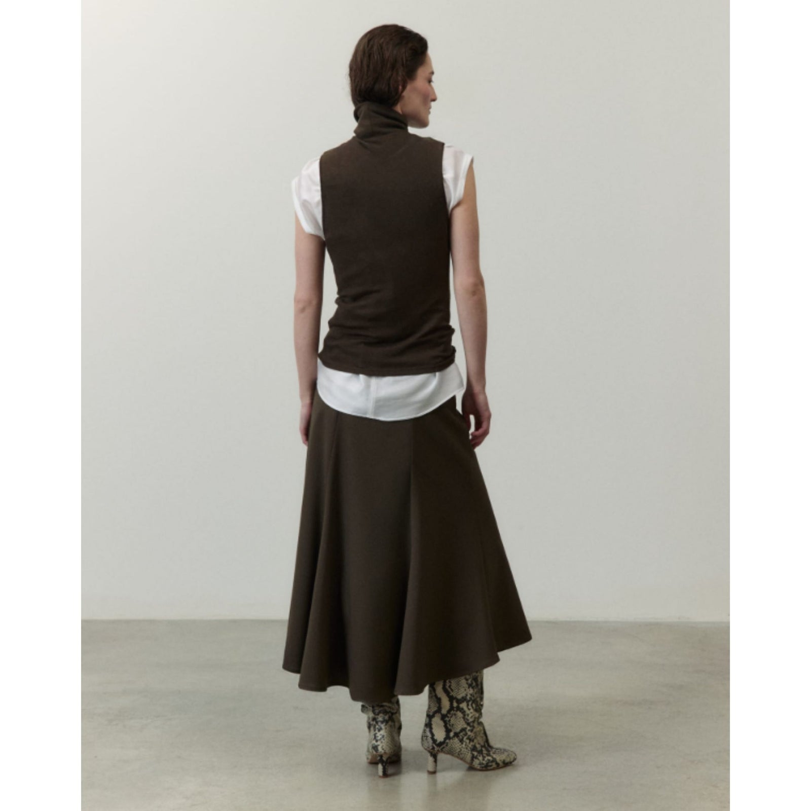 A model stands in a minimalist studio setting wearing a dark brown sleeveless mock-neck top over a white turtleneck, paired with a matching dark brown midi skirt and snakeskin-patterned boots.