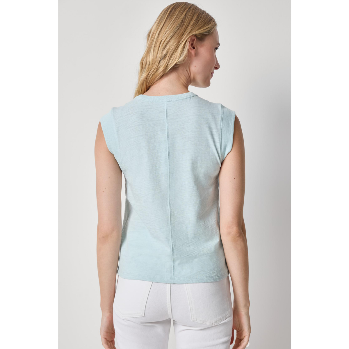 Woman wearing a light blue sleeveless top and white pants on a plain background