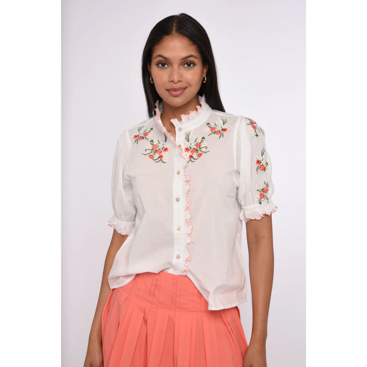 Woman wearing a white blouse with floral embroidery and coral pants on a white background
