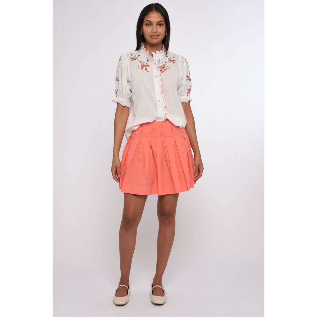 Woman wearing a white blouse with floral embroidery and a coral skirt on a white background