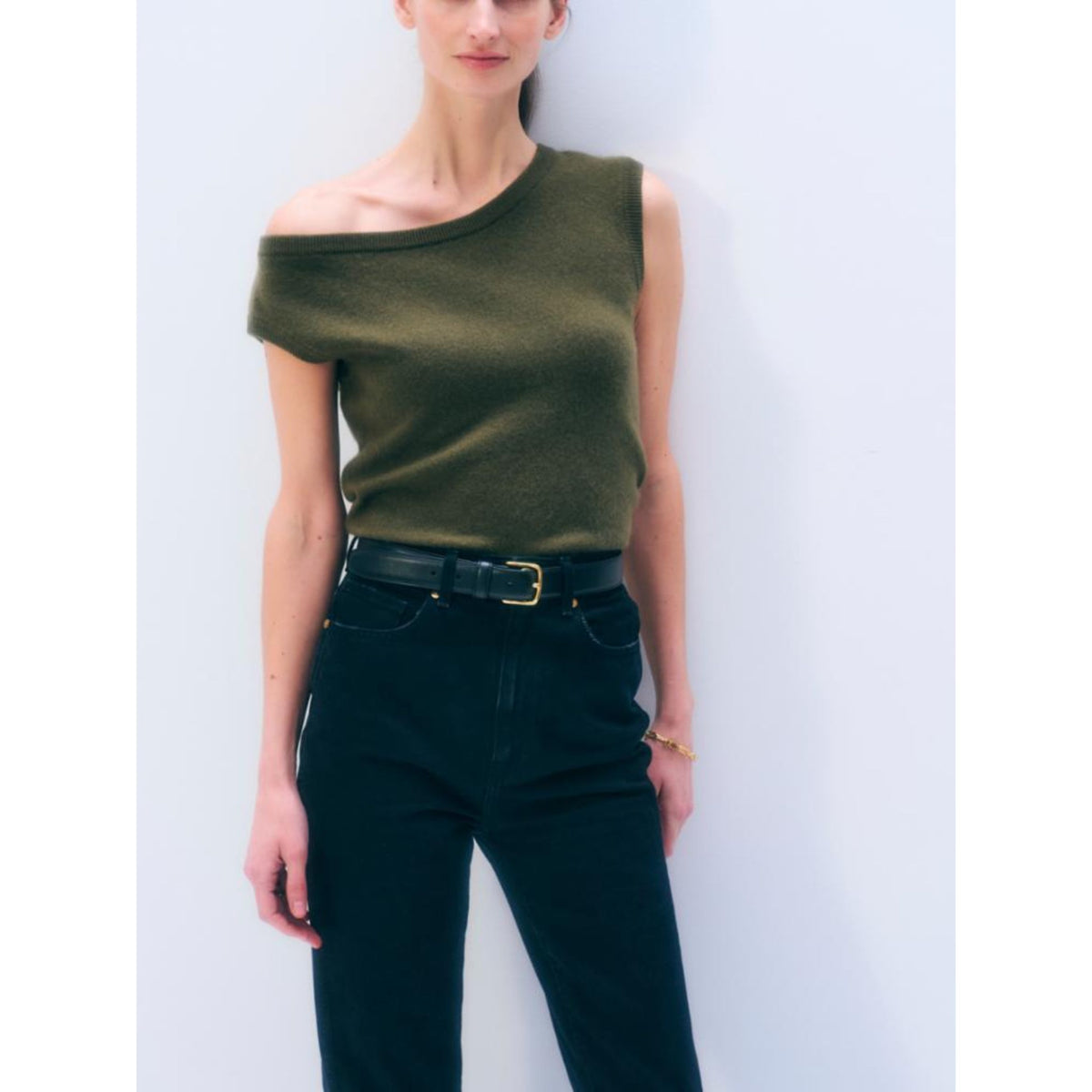 Front view of a model wearing the short-sleeve "Cashmere Off Shoulder" top in Olive green with an asymmetric off-the-shoulder neckline, styled with dark denim jeans and a black belt.