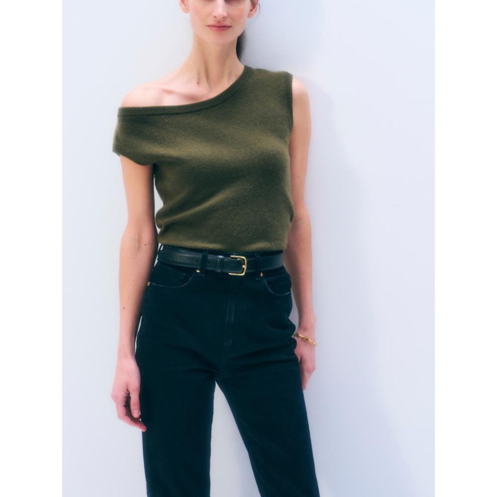 Front view of a model wearing the short-sleeve "Cashmere Off Shoulder" top in Olive green with an asymmetric off-the-shoulder neckline, styled with dark denim jeans and a black belt.