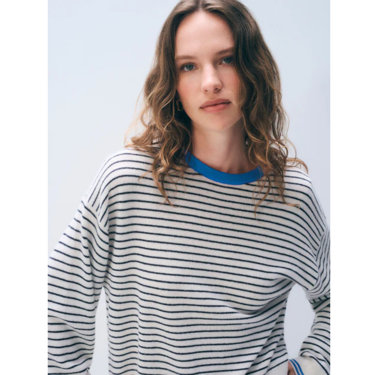 A woman wearing a long-sleeve knit top with thin black horizontal stripes on a white or light gray base, featuring a contrasting bright blue crew neck.