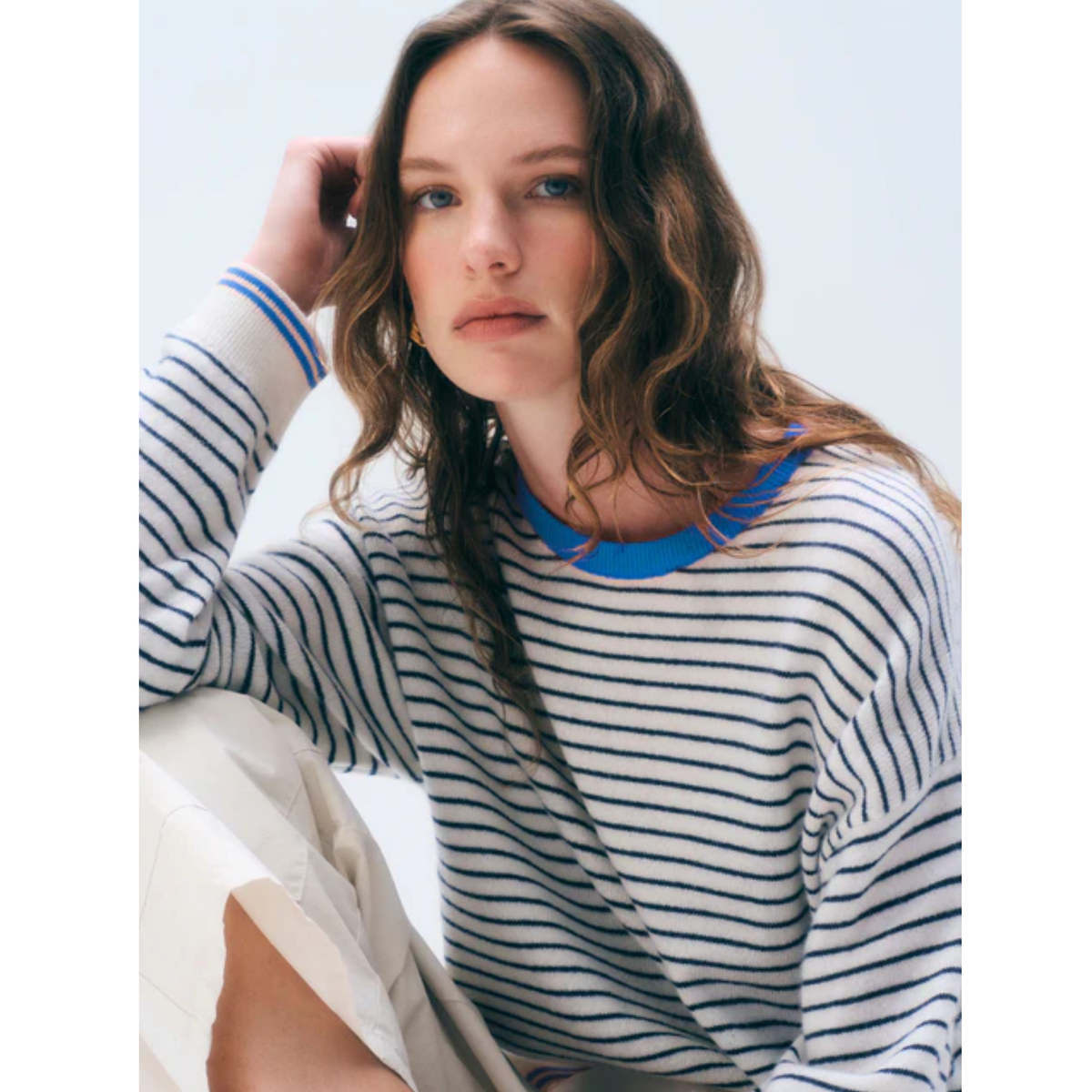 A woman wearing a long-sleeve crewneck top with thin navy horizontal stripes on a white base, featuring a bright blue collar trim, paired with light beige trousers.
