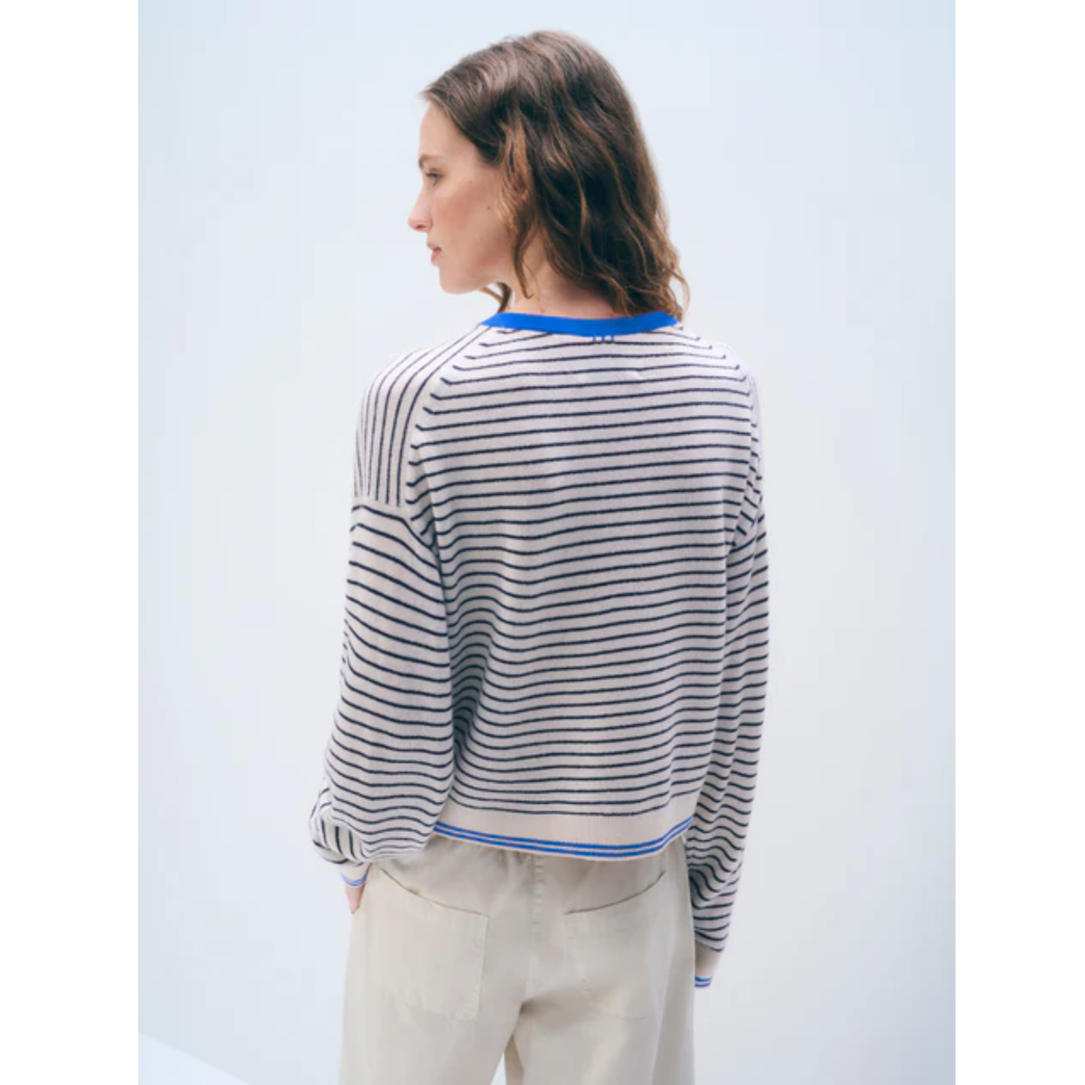 A woman shown from the back wearing a cropped, long-sleeve knit sweater with thin black horizontal stripes on a white background, featuring blue ribbed trim at the collar and hem, paired with light beige trousers.