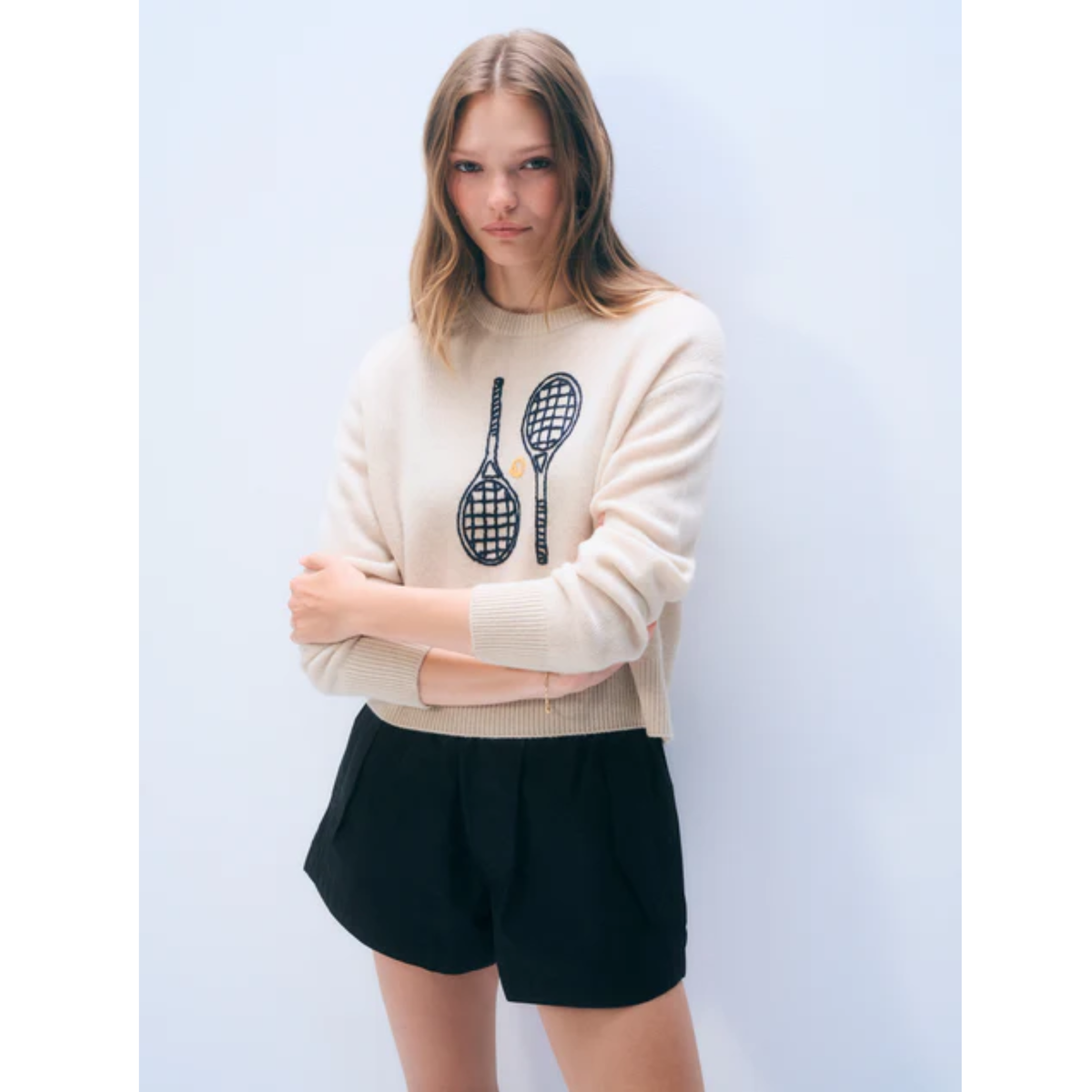 A woman modeling a cream-colored knit sweater with a black tennis racket graphic on the front, paired with black shorts, set against a light blue studio background.