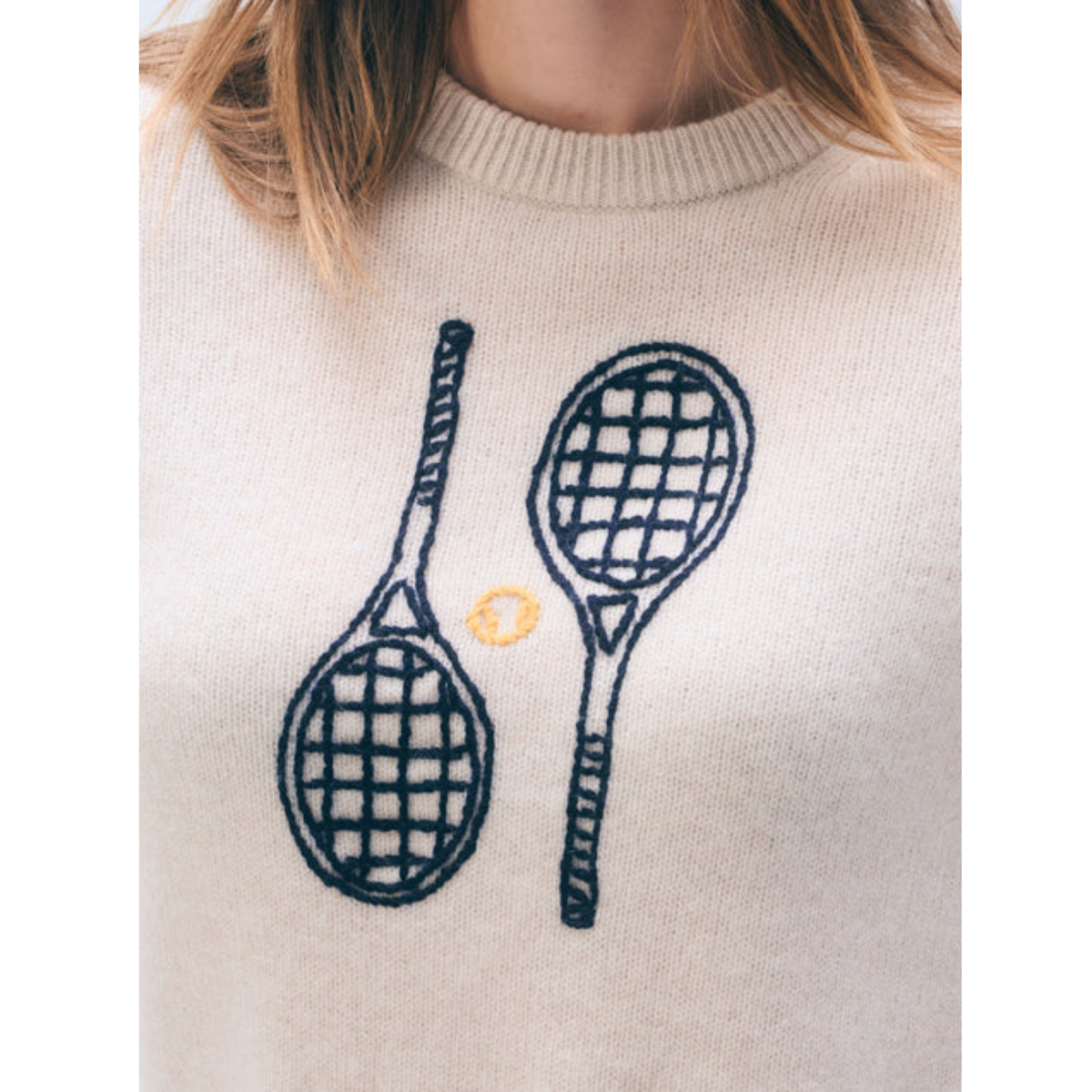 A close-up of a cream-colored knit sweater featuring a graphic of two tennis rackets and a small yellow tennis ball on the front.