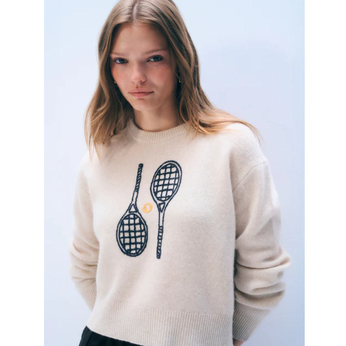 A woman wearing a cream-colored knit sweater with a graphic of two black tennis rackets and a small yellow tennis ball on the front, set against a light blue background.