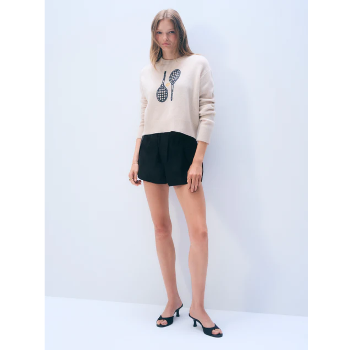 A woman modeling a beige knit sweater with a black tennis racket graphic on the chest, paired with black shorts and black mule sandals, set against a plain light gray studio background.
