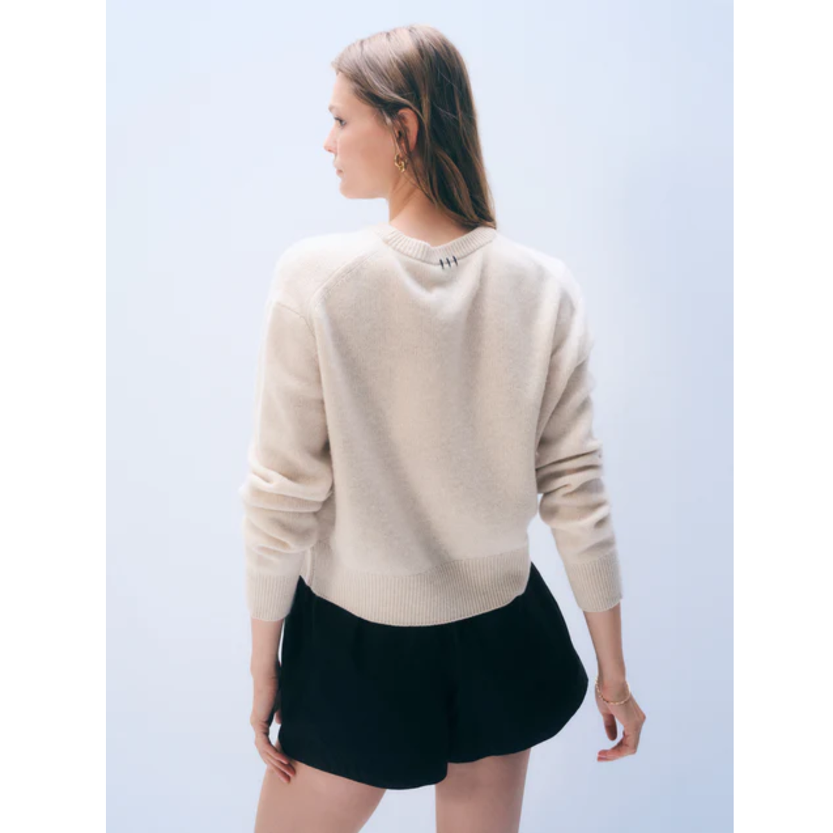 A woman shown from the back wearing a light cream knit sweater and black shorts against a pale blue studio background.