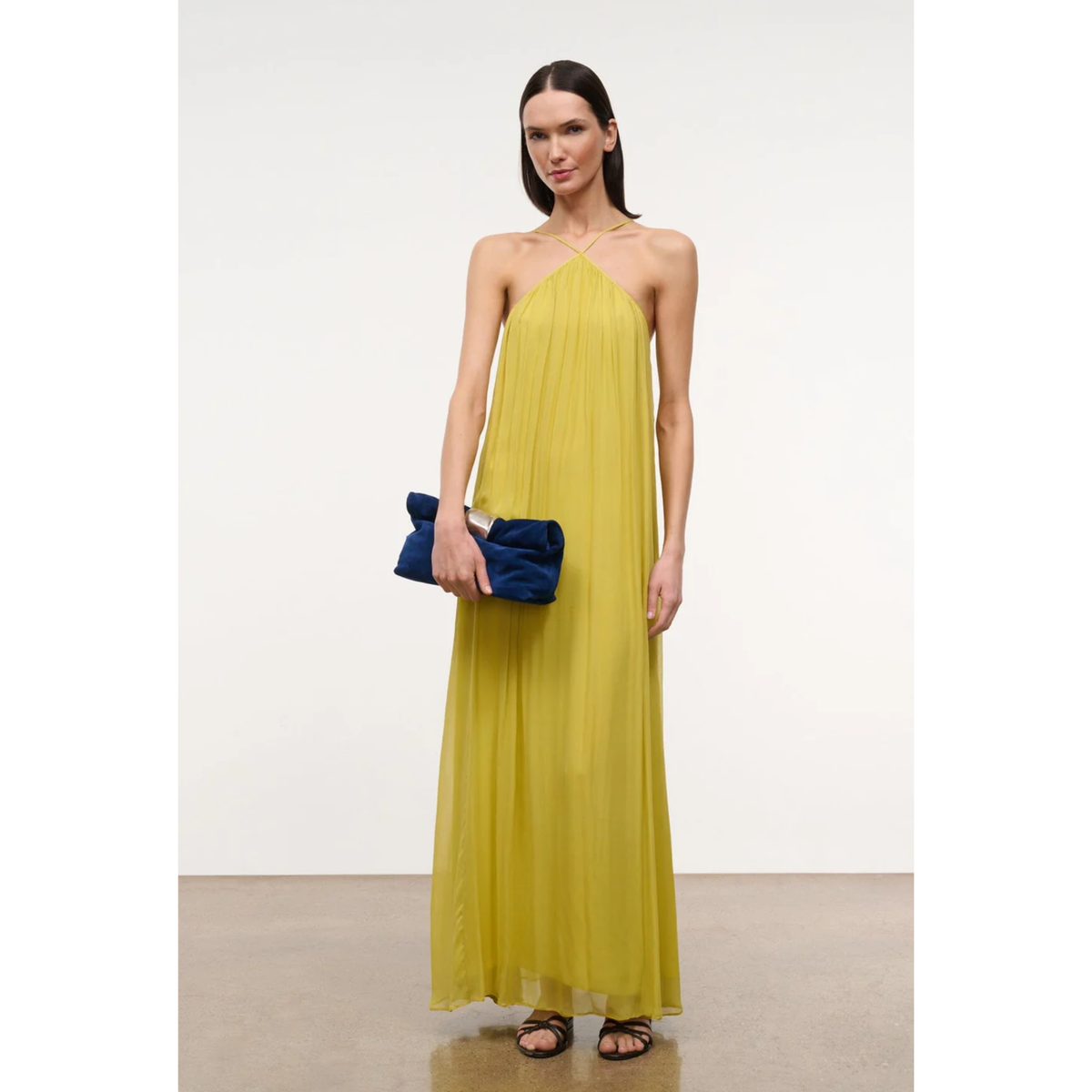 A woman modeling a sleeveless, halter-neck, floor-length yellow dress, holding a dark blue clutch purse against a plain off-white studio background.