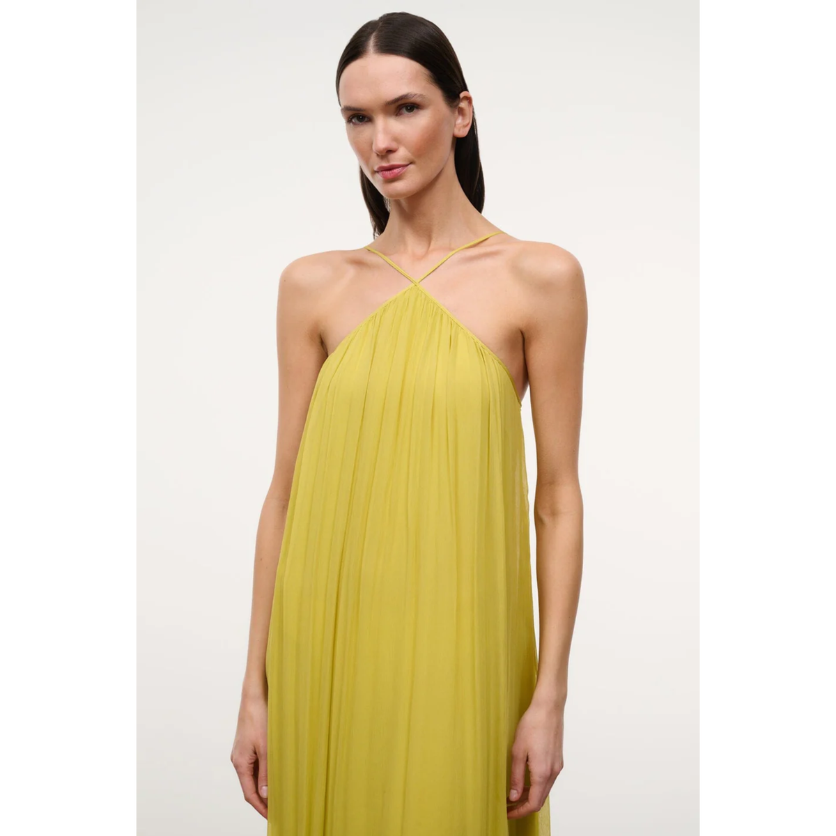 A woman modeling a sleeveless, pleated halter-neck dress in a vibrant yellow against a plain light gray studio background.