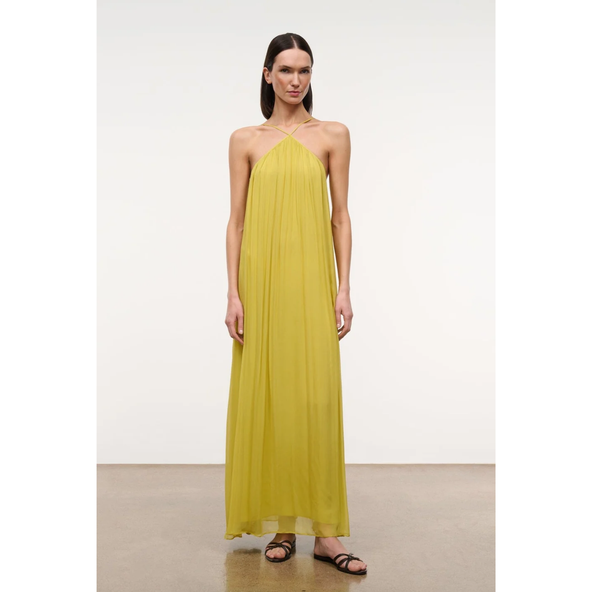 A woman modeling a sleeveless, halter-neck, floor-length yellow dress with a flowing, pleated fabric, set against a minimalist studio background.