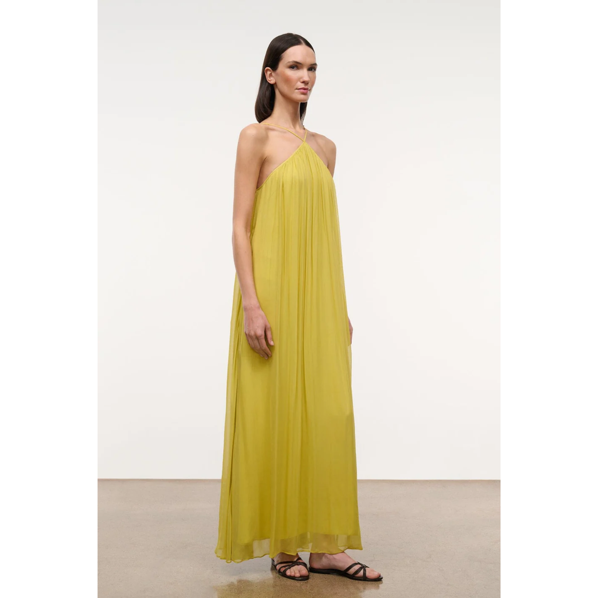 A woman modeling a flowing, sleeveless halter-neck maxi dress in a vibrant yellow, paired with black strappy flat sandals, set against a minimalist off-white studio background.