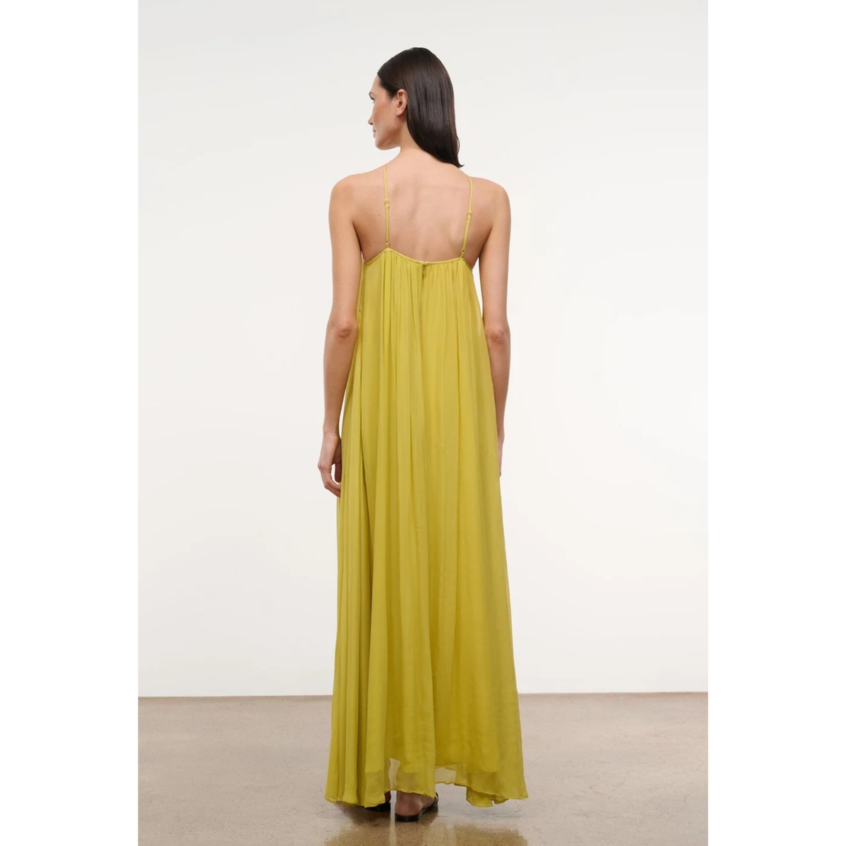 A woman wearing a flowing yellow maxi dress, shown from the back in a studio setting with a plain white wall and light floor.