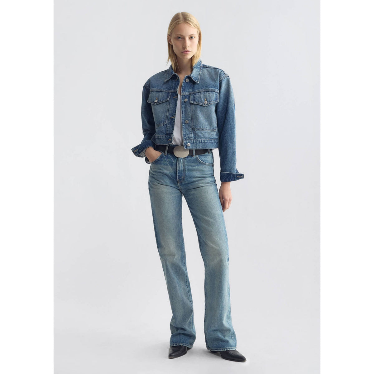 Full-length front view of a model posing in the cropped blue denim jacket paired with matching double denim jeans.