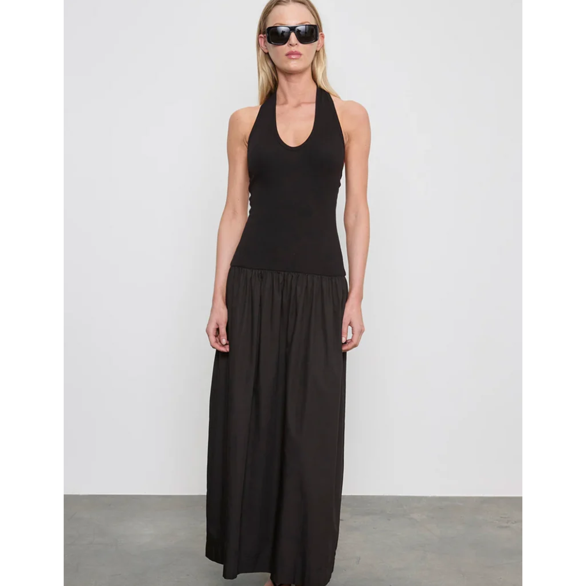 A woman modeling a black sleeveless halter-neck maxi dress with a gathered waist, standing against a light gray studio background.