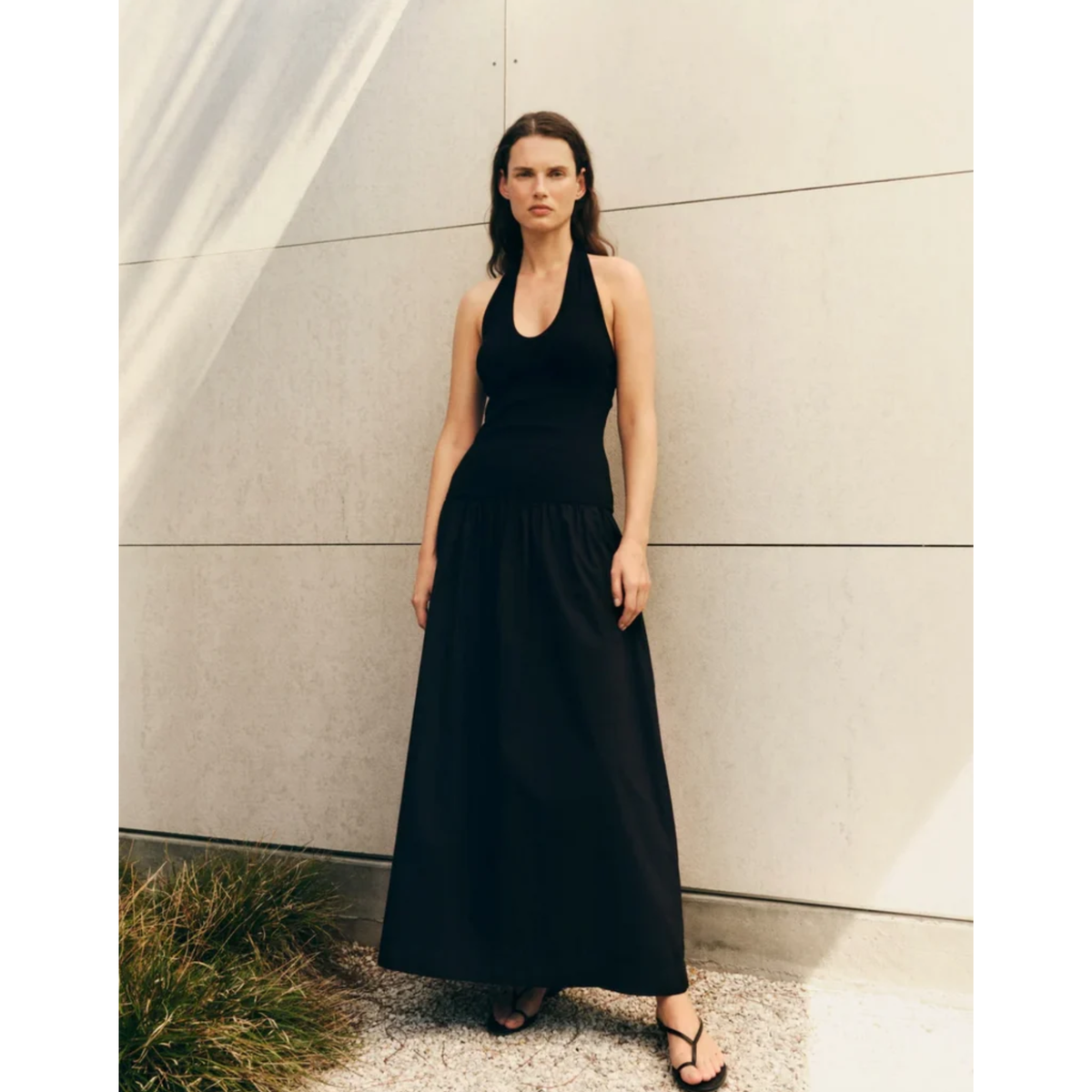 A woman stands outdoors against a light stone wall wearing a sleeveless black halter maxi dress and black sandals.