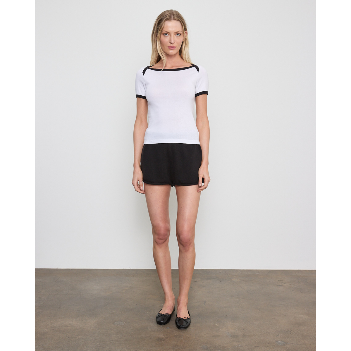 A woman stands in a studio wearing a white short-sleeve top with black trim and black shorts, paired with black flats.