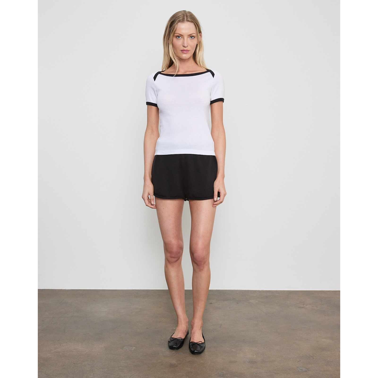 A woman stands in a studio wearing a white short-sleeve top with black trim and black shorts, paired with black flats.