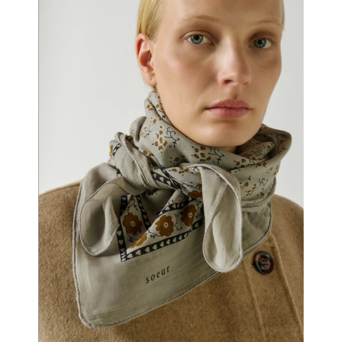 A close-up of a model wearing a beige scarf with a brown and black floral pattern and a tan coat. The scarf has the brand name 'soeur' printed on it.