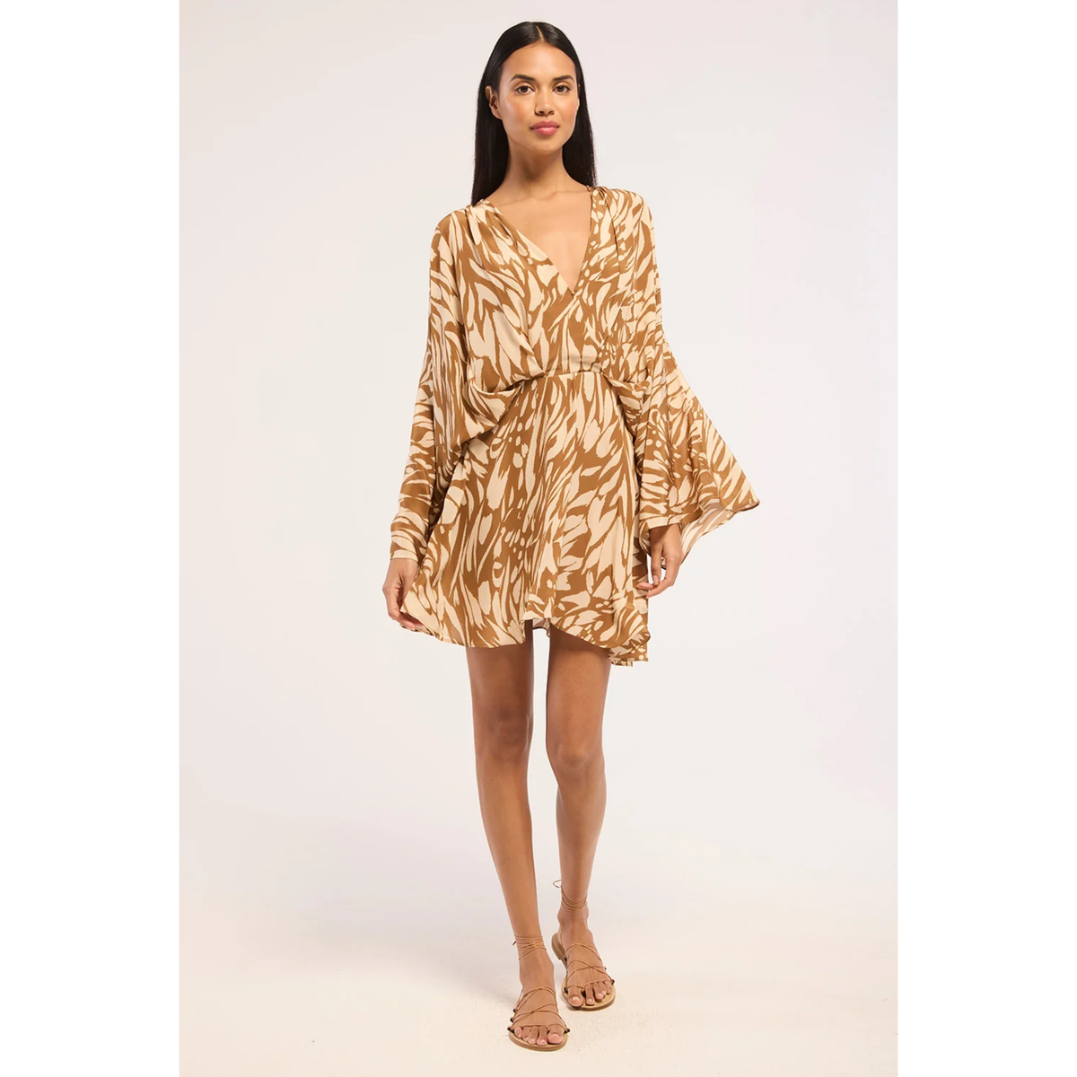 A woman modeling a long-sleeve mini dress with a brown and beige leafy pattern, a wrap-style V-neckline, and flared bell sleeves, photographed against a plain light background.