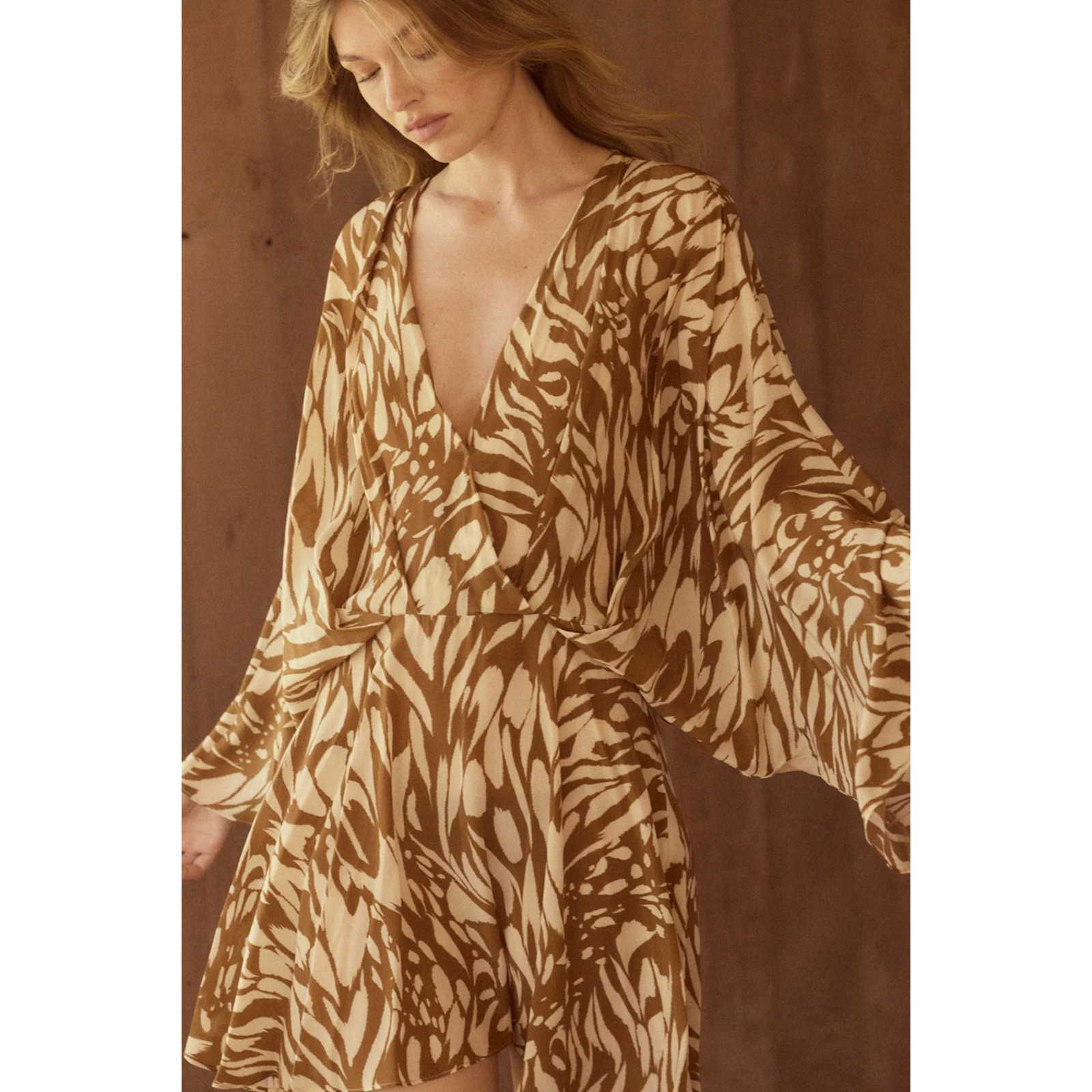 A woman wearing a beige and brown abstract print wrap dress with long flowing sleeves, photographed against a warm brown wood-textured background.