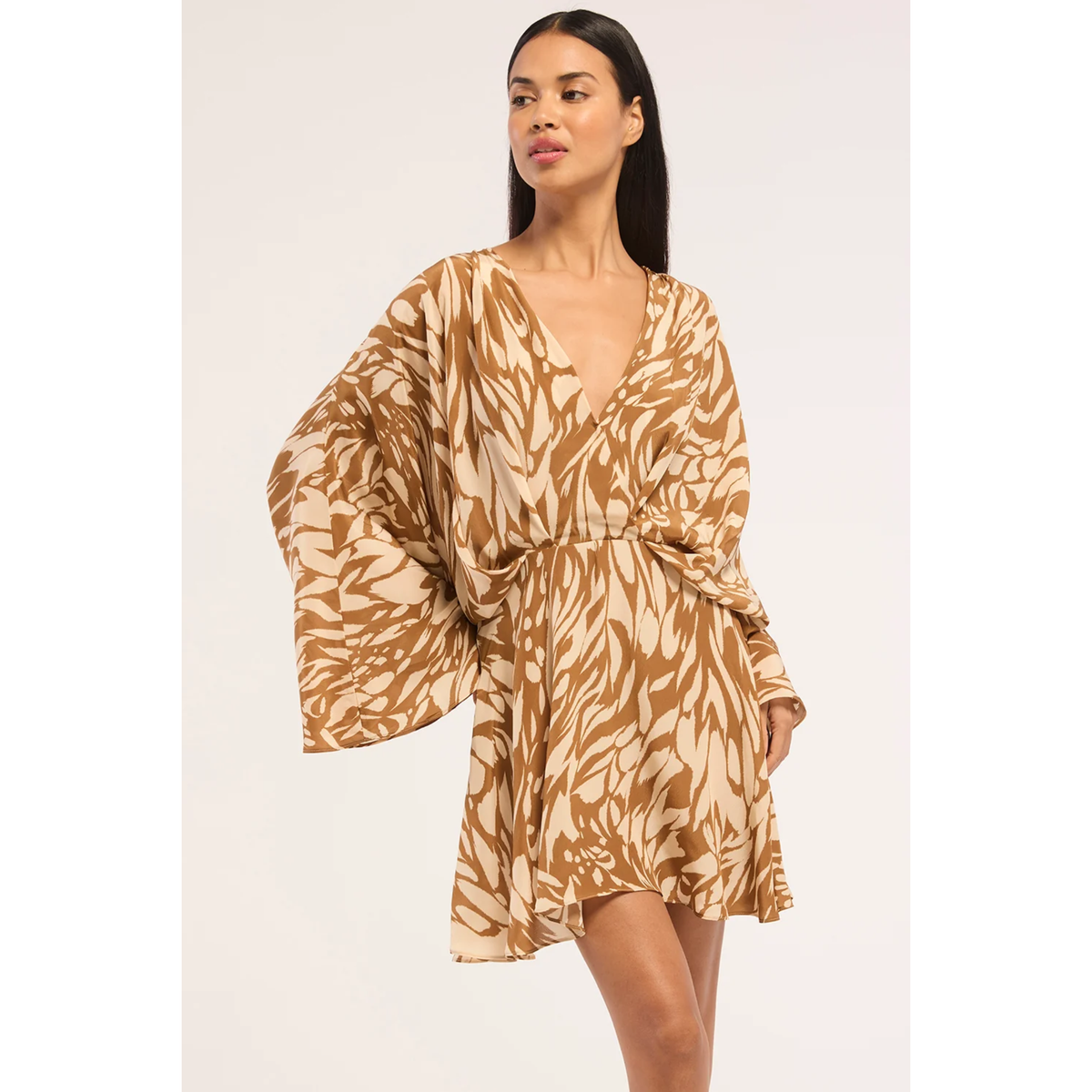 A woman modeling a short, loose-fitting dress with wide sleeves and a tied waist, featuring a tan and cream abstract leaf-like print against a plain light background.