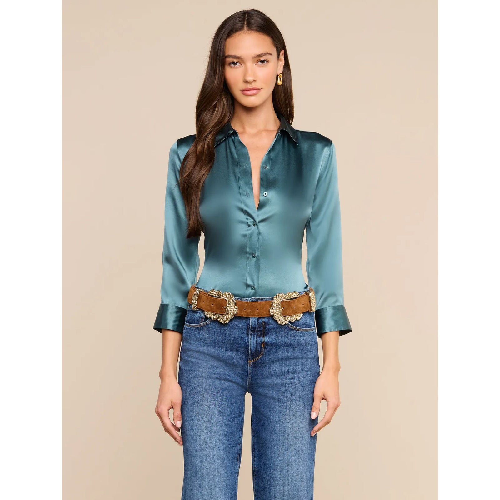 A woman modeling a satin long-sleeve button-up blouse in a teal blue-green color, paired with blue jeans and a brown western-style belt with a large gold buckle, against a solid beige background.