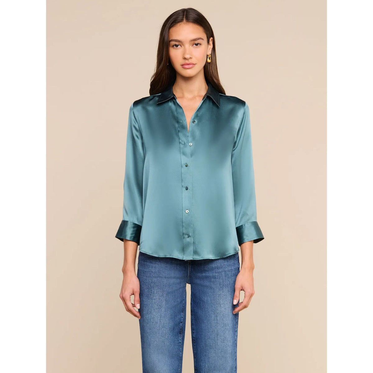 A woman modeling a satin button-up blouse in a muted teal-green color with matching satin-trimmed cuffs, paired with blue jeans, against a solid beige background.