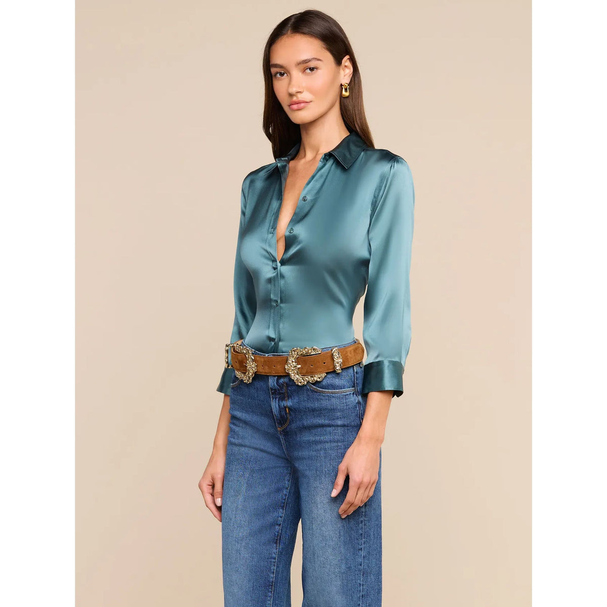 A woman modeling a satin button-down blouse in a muted teal-green color, paired with blue jeans and a brown western-style belt, against a solid beige background.
