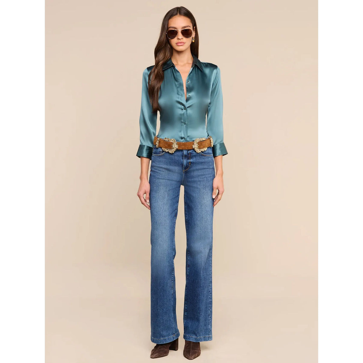 A woman modeling a satin-like teal button-up blouse with three-quarter sleeves, paired with blue jeans, a brown belt, and sunglasses against a plain beige studio background.
