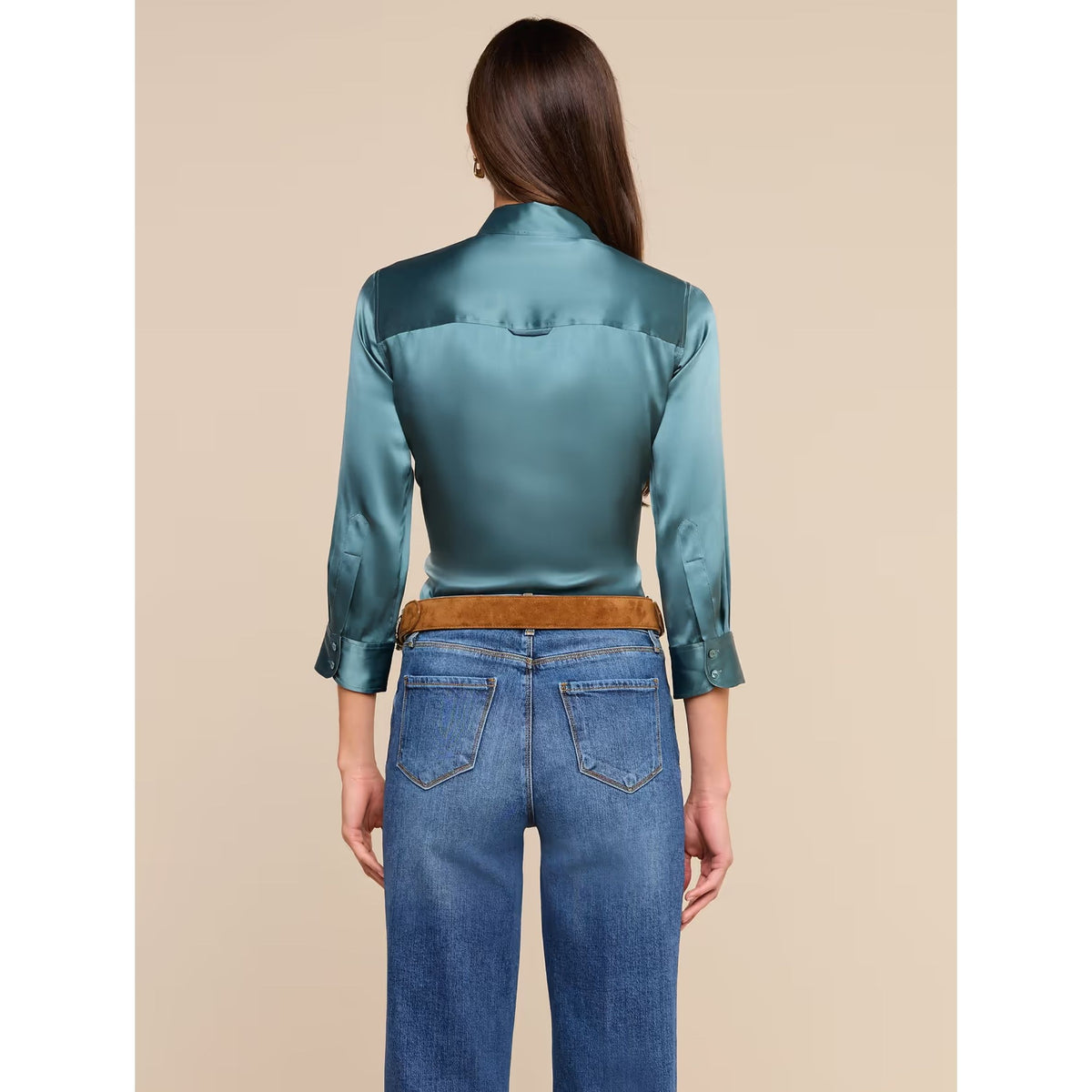 A woman shown from the back wearing a teal satin button-up shirt tucked into blue jeans with a brown belt, against a solid beige background.