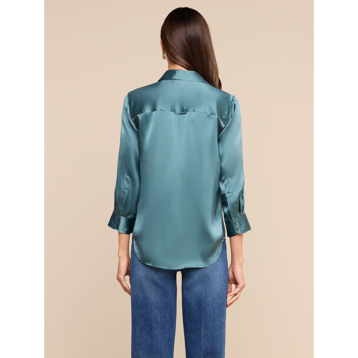 A woman shown from the back wearing a satin-like teal blouse with three-quarter sleeves and blue jeans, standing against a plain beige studio background.