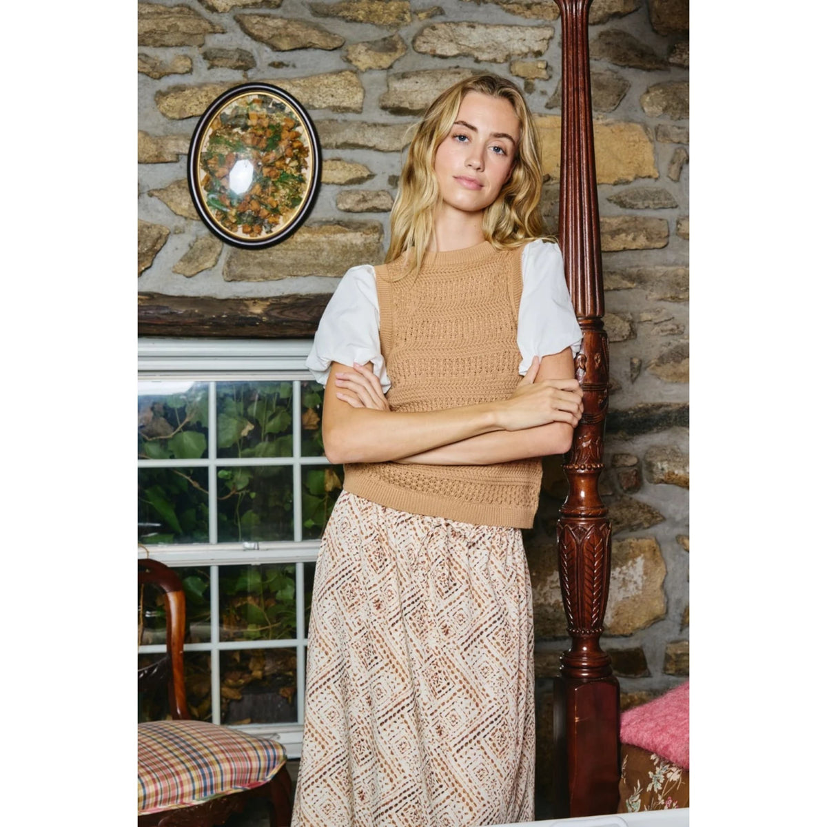 A woman stands indoors against a rustic stone wall, wearing a tan sleeveless knit top with white puffed short sleeves and a beige patterned skirt. She is posed beside a dark wooden bed frame, with a window and wall decor visible in the background.